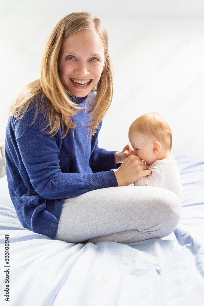 Mother playing with child and smiling at camera. Happy young mother with adorable infant daughter playing together in bedroom. Family concept