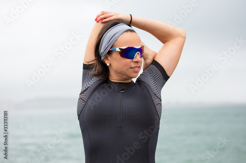 Woman stretching on sea coast. Sporty middle aged woman in sportswear and goggles stretching arms near sea during daytime. Triathlon concept