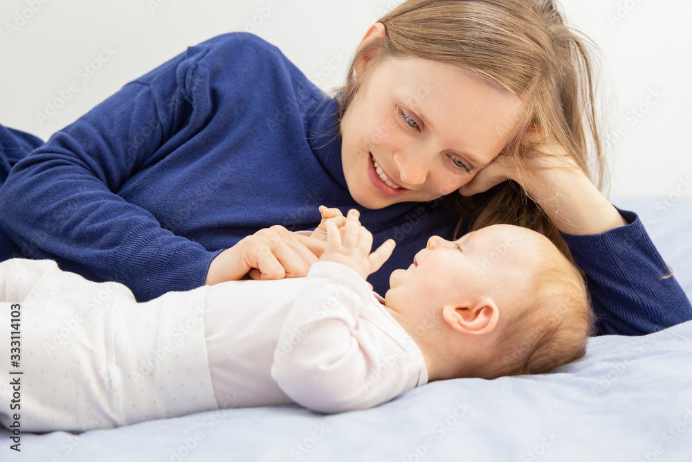 Young mother playing with cute baby on bed. Happy young mother looking at adorable little daughter lying on bed. Motherhood concept Young mother playing with cute baby on bed. Happy young mother looking at adorable little daughter lying on bed. Motherhood concept