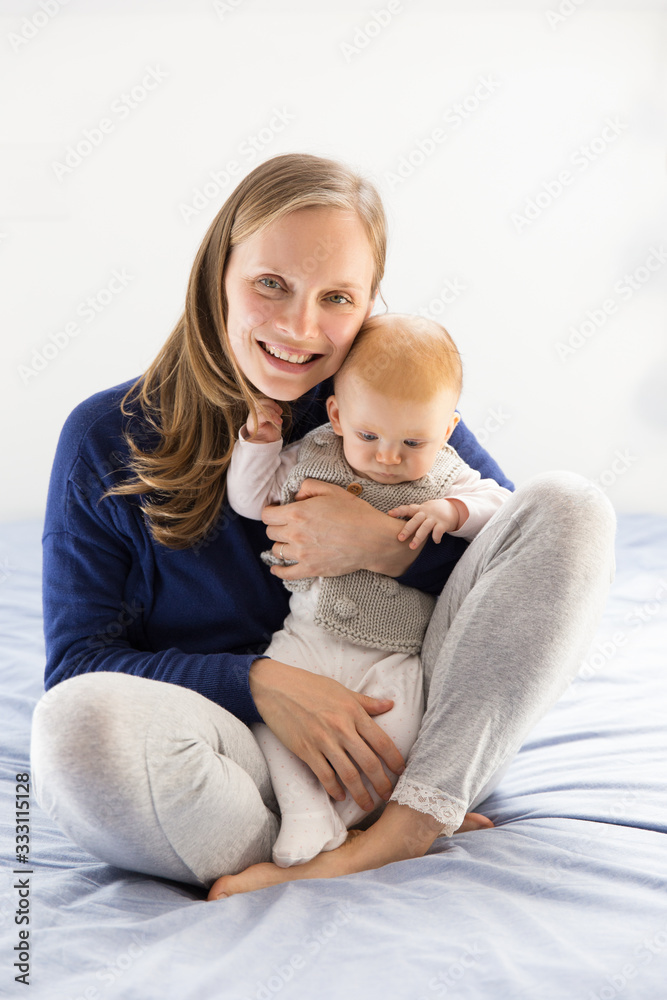 Positive smiling new mom cuddling adorable little baby girl. Young woman nursing six month daughter in bedroom at home. Motherhood concept Positive smiling new mom cuddling adorable little baby girl. Young woman nursing six month daughter in bedroom at home. Motherhood concept
