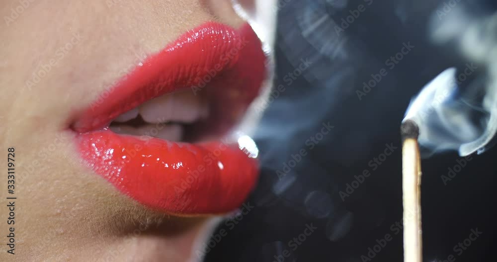 Detail close-up macro shot of the lips of a girl painted with a shiny red lipstick  blowing out the fire of a burning match on black studio background isolated. 4k slow motion 50 fps