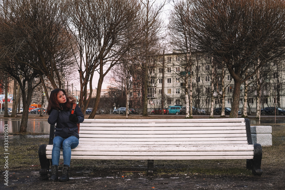 A brunette girl with long hair wearing a jacket and carrying a backpack is sitting on a white bench in the middle of the Park in spring