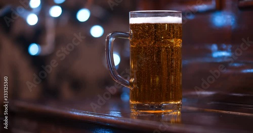 Close up of one half liter glass mug full of beer standing on a brown varnished wooden bar counter in a dark pub with blinking lights in the background. With free space for writing 4K