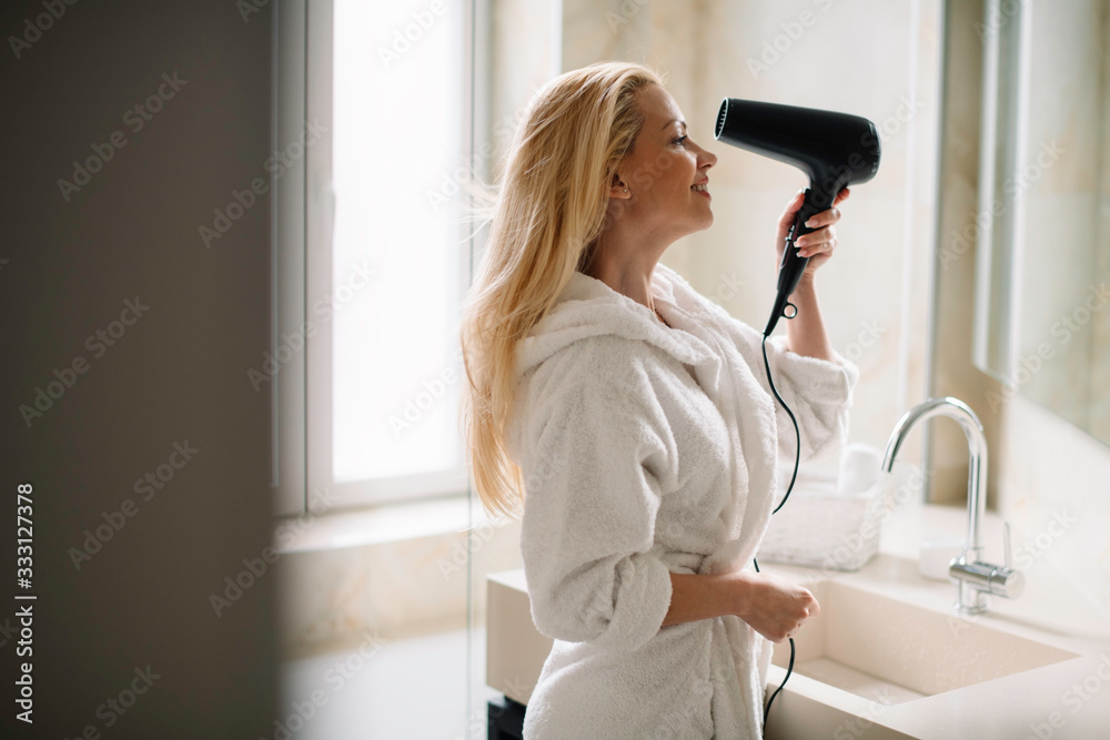 Young happy smiling woman wearing bathrobe using hair dryer in bathroom.
