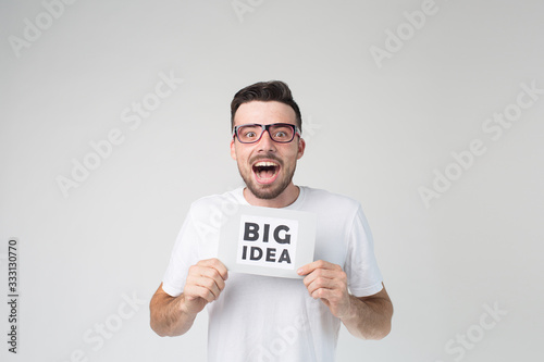 Young man isolated over background. Guy has a big idea. Hold piece of paper with hnds and smile. Look straight through glasses. Exited and emotional happy.