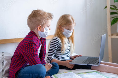 Distance learning online education. Sickness school boy and girl in medical mask studying at home with digital tablet laptop notebook and doing homework. Sitting on bed with training books.