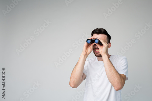 Young man isolated over background. Guy look at binoculars far away. Hold with both hands and investigate. Spy or casual looking guy.