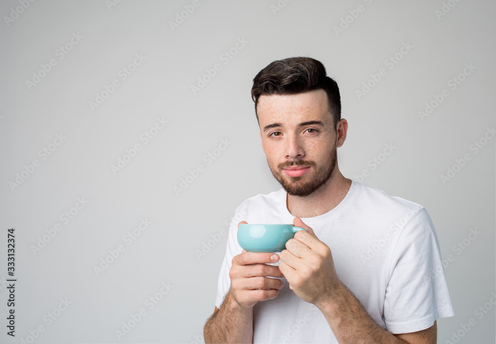 Young man isolated over background. Casual guy in white shirt hold blue cup in hands with tea or coffee inside. Loook straight on camera and pose. Ready to drink warm or hot drink. Young man isolated over background. Casual guy in white shirt hold blue cup in hands with tea or coffee inside. Loook straight on camera and pose. Ready to drink warm or hot drink.