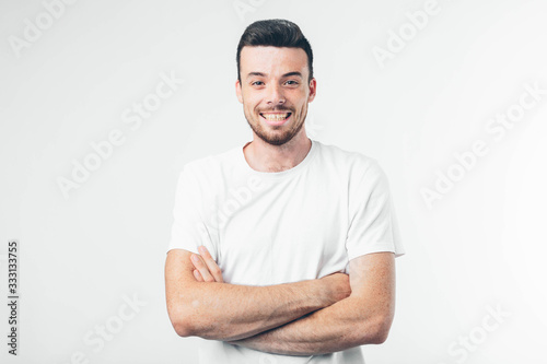 Young brunette man with beard dressed in white T-shirt. Guy confident and wide smiling. Arms crossed at chest level. Isolated over light background.