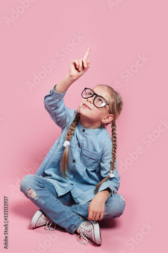 child with glasses points finger up on pink background
