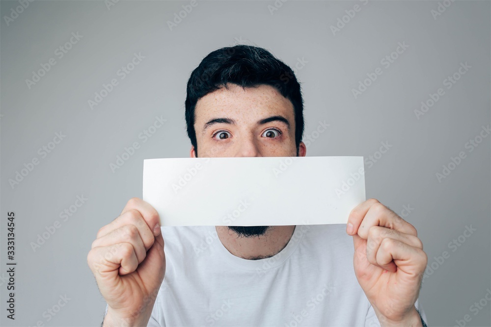 Young man isolated over background. Portrait of guy in white shirt with windered sight. Cover mouth and lips with piece of white page Young man isolated over background. Portrait of guy in white shirt with windered sight. Cover mouth and lips with piece of white page
