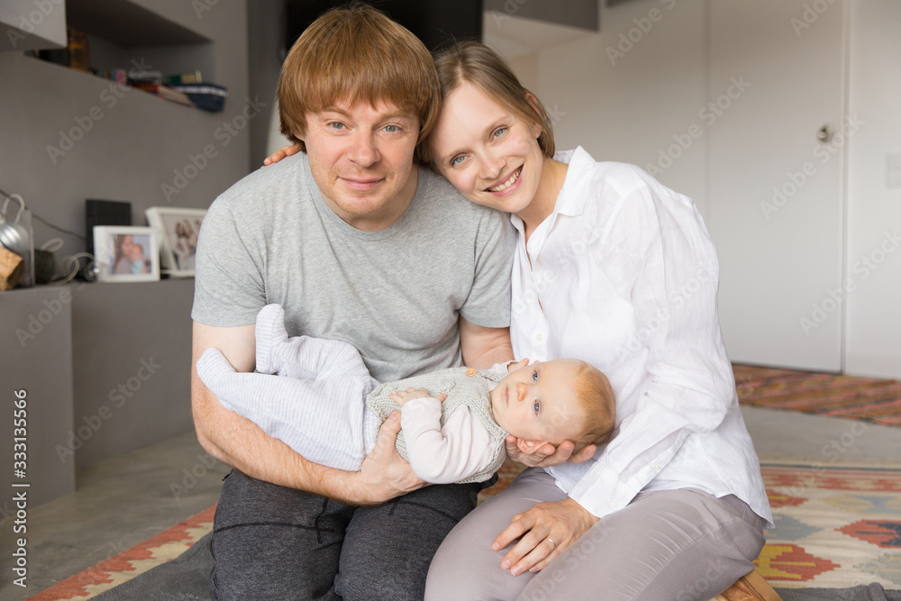 Cheerful positive new parents nursing baby daughter together. Young couple holding six month child and looking at camera. Parenthood concept Cheerful positive new parents nursing baby daughter together. Young couple holding six month child and looking at camera. Parenthood concept