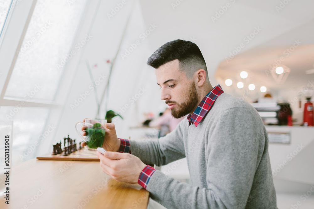Young bearded man in cafe. He dressed in gray pullover and plaid shirt. Man holds tea and looks at smartphone . Chessboard flower and blurred interior of cafe on background. Young bearded man in cafe. He dressed in gray pullover and plaid shirt. Man holds tea and looks at smartphone . Chessboard flower and blurred interior of cafe on background.