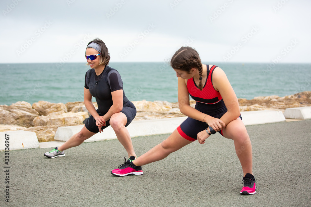 Athletic women stretching near sea. Full length view of sporty women in sportswear stretching and working out on sea coast. Triathlon concept Athletic women stretching near sea. Full length view of sporty women in sportswear stretching and working out on sea coast. Triathlon concept