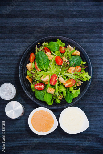 Bowl of healthy green vegetable salad on top with tomatos and biscuits. Side dish with salt pepper bottle, white cream and thousand island salad dressing
