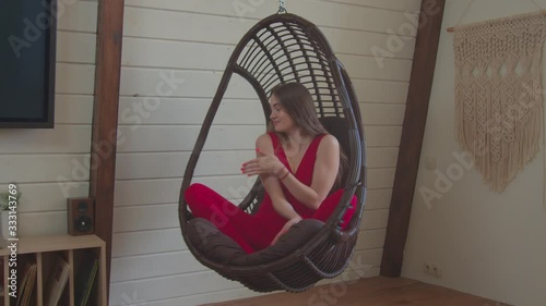 Portrait of gorgeous elegant young woman with beautiful long straight hair in stylish red sports overalls enjoying leisure in hanging chair at home, expressing modest allure and wellness.