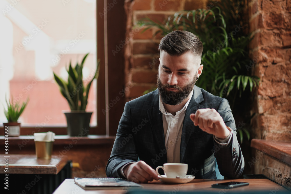 A young handsome stylish bearded man Kavkuzets chdmt at a table in the restaurant opposite the window. He reads a magazine, drinks coffee. A young handsome stylish bearded man Kavkuzets chdmt at a table in the restaurant opposite the window. He reads a magazine, drinks coffee.