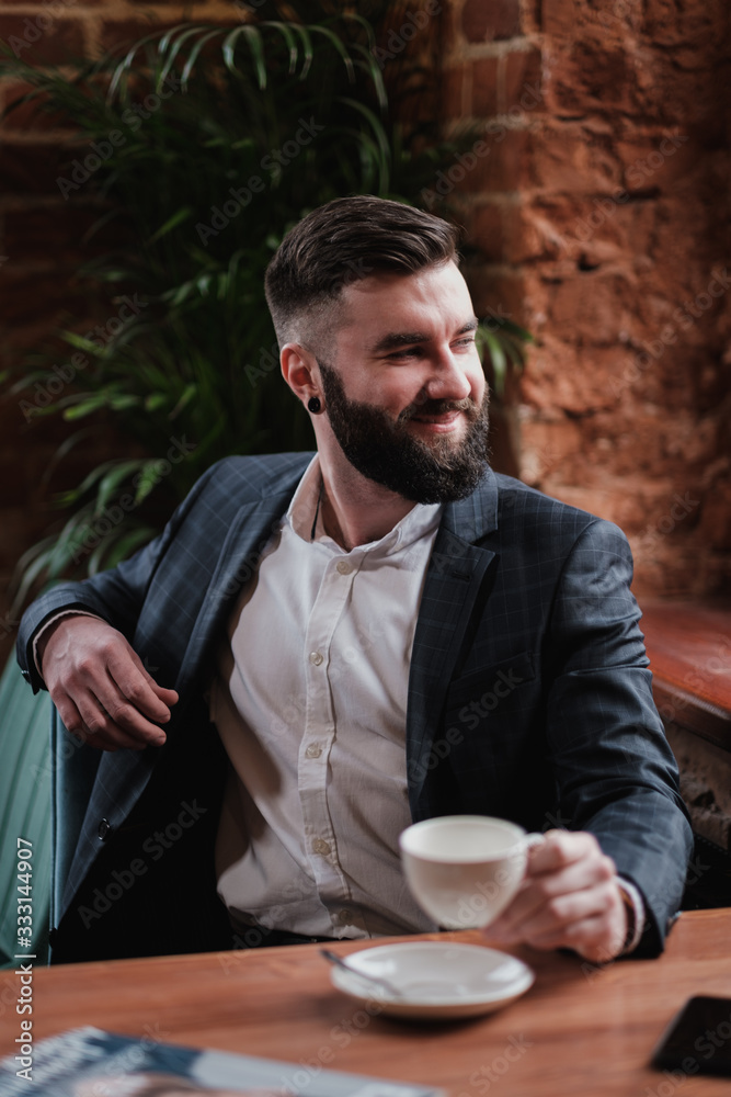 A young handsome stylish bearded man Kavkuzets chdmt at a table in the restaurant opposite the window. He reads a magazine, drinks coffee. A young handsome stylish bearded man Kavkuzets chdmt at a table in the restaurant opposite the window. He reads a magazine, drinks coffee.