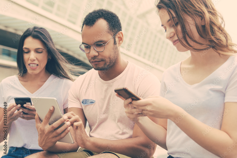 Group of young people using devices on street. Calm friends sitting with smartphones and tablet. Technology concept Group of young people using devices on street. Calm friends sitting with smartphones and tablet. Technology concept