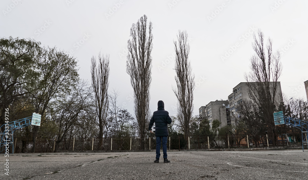 concept of epidemic and quarantine - a boy with a face mask and a ball alone on the sports area in the city concept of epidemic and quarantine - a boy with a face mask and a ball alone on the sports area in the city
