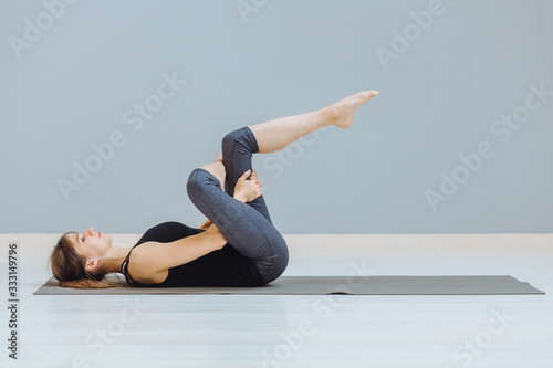 Relaxing back pain exercise concept. Attractive sportive woman doing pilates exercise lying on yoga mat at empty room at grey wall ackground.