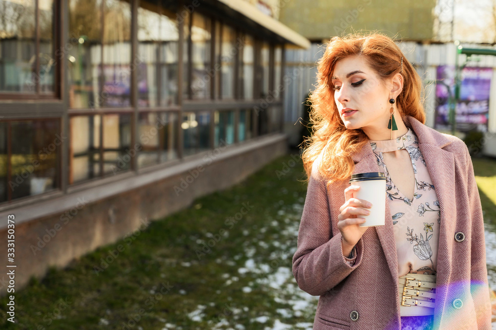 Photo of a fashionable beautiful woman with long hair in the spring city in the sunshine outdoors with a cup of coffee in her hands