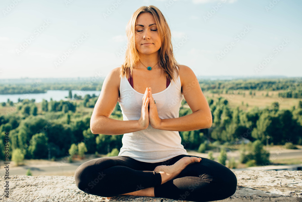 Young woman practices yoga outside. Calm smiling girl sitting on parapet in lotus position with close eyes. Her hands at chest level touch each other. Trees river and sky on background.