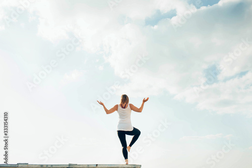 Young woman practices yoga outside. Blonde girl turned back standing on one leg, second raised bent at knee. Hands on sides bent at elbows and turned up. Sky and clouds on background.
