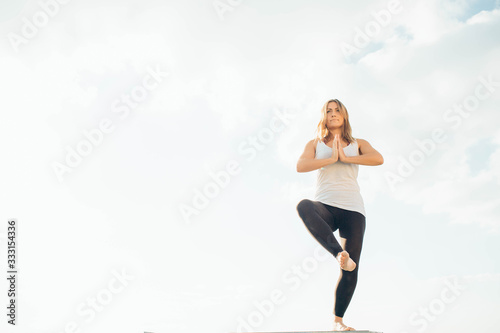 Young woman practices yoga outside. Blonde girl standing on one leg, another raised bent at knee. Hands at chest level, palms touching. Sky and clouds on background.