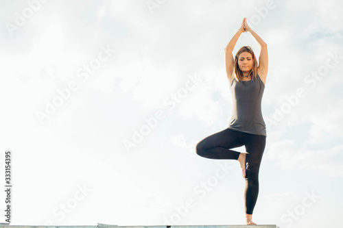 Young woman practices yoga outside. Blonde girl standing on parapet do tree pose. Hands above head touching. She dressed in black leggings and grey T-shirt. Sky and clouds on background.