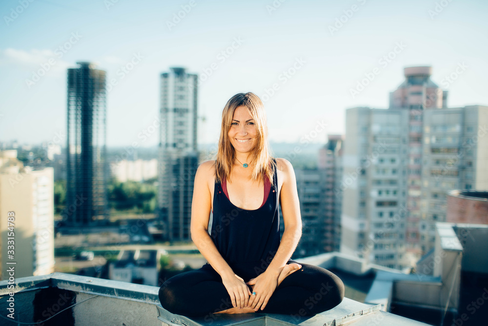 Fototapeta premium Young woman practices yoga outside. Calm smiling girl sitting on parapet in lotus position with open eyes. Her hands lowered to ankles. City and high-rise buildings on background.