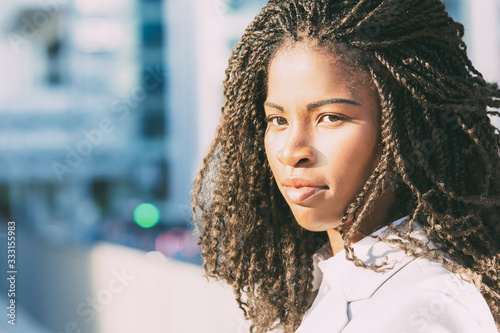 Pensive beautiful student girl posing in city. Closeup of young African American woman with dreads standing outside and looking at camera. Female portrait concept
