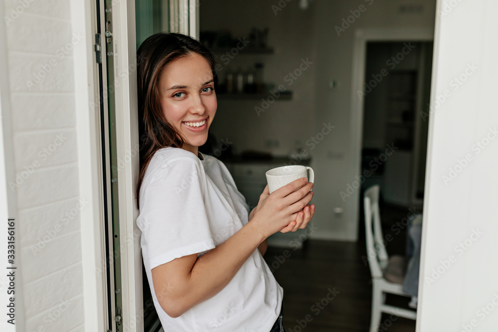 Portrait of young european woman with dark hair and healthy skin smiling at camera with morning coffee in the modern light kitchen Portrait of young european woman with dark hair and healthy skin smiling at camera with morning coffee in the modern light kitchen