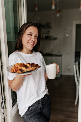 Caucasian smiling woman wearing white t-shirt with dark hair and healthy skin holding morning coffee and croissants for breakfast at home