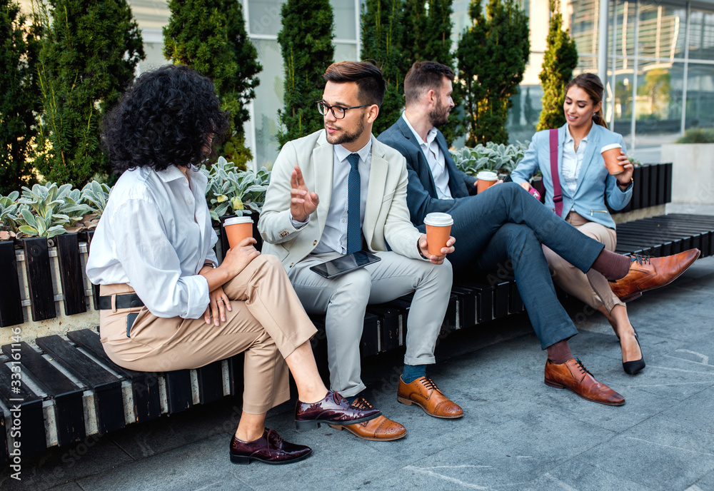 Fototapeta premium Group of coworkers having a coffee brake together, siting outside in front of office buildings and talking.