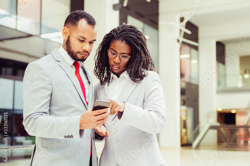 Focused diverse colleagues using mobile phone together. Business man and woman wearing formal suits, holding smartphone and pointing at screen. Watching content concept