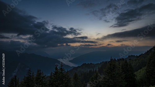  Dark forest after sunset with clouds and lake in background. Switzerland