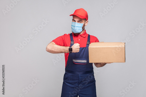 Delivery on quarantine. Ontime service! man with surgical medical mask in blue uniform and red t-shirt standing, holding delivery box and showing watch on grey background. Indoor shot, isolated, photo