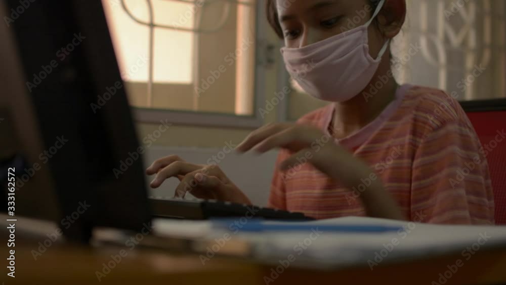 Cute girl wearing protective mask working with computer and writing on the desk at home during COVID-19 outbreak situation. Study at home concept. Cute girl wearing protective mask working with computer and writing on the desk at home during COVID-19 outbreak situation. Study at home concept.