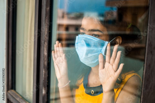 Coronavirus. Sick young girl of corona virus looking through the window and wearing mask protection and recovery from the illness in home. Quarantine. Patient isolated in house to prevent infection.