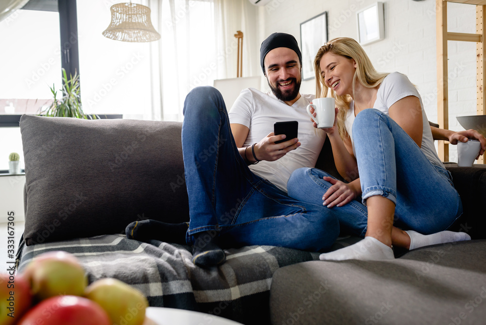 Obraz premium Young hipster couple sitting on sofa at home talking, drinking coffee, looking at smartphone