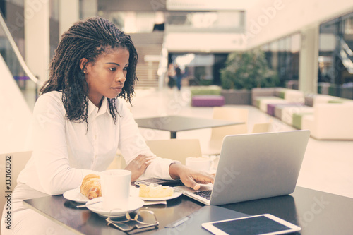 Serious professional watching presentation on laptop during lunch. Young African American business woman drinking coffee in cafe, using computer. Watching content concept © Mangostar