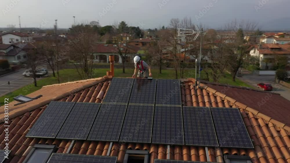 Aerial circle orbit shot of technician with hard hat and safety equipment working on maintenance of photovoltaic panel system installed on home domestic roof top, urban landscape. Wide angle camera.