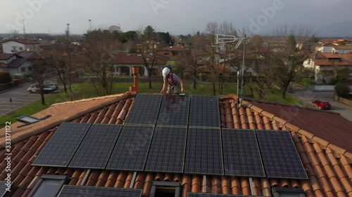 Aerial circle orbit shot of technician with hard hat and safety equipment working on maintenance of photovoltaic panel system installed on home domestic roof top, urban landscape. Wide angle camera.