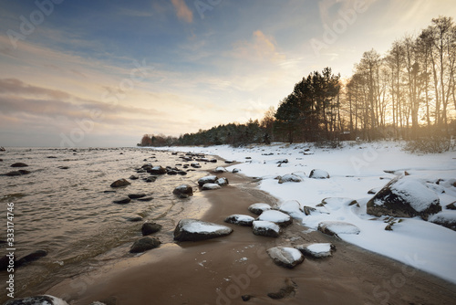 A view of the snow-covered Baltic sea coast at sunset. Stones in the water close-up. Coniferous forest in the background. Stunning cloudscape. Warm evening light. Kaltene, Latvia photo