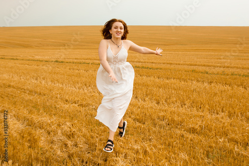 Runs across the field. Young beautiful redhead woman in the middle of a wheat field having fun. Summer landscape, good weather. Windy day with the sun and clouds. White cotton dress, eco style. © Yuliia