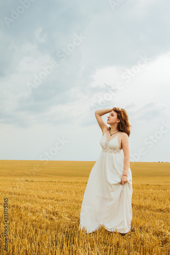 Good outdoor recreation. Young beautiful redhead woman in the middle of a wheat field, having fun. Summer landscape, good weather. Windy day with the sun and clouds. White cotton dress, eco style.