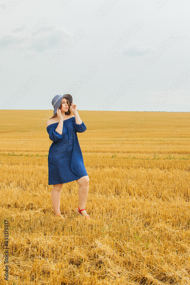 Young beautiful blonde girl in the middle of a wheat field. Posing looking at the camera. Summer landscape, good weather. Blue cotton dress, eco style. Beautiful striped hat. Young beautiful blonde girl in the middle of a wheat field. Posing looking at the camera. Summer landscape, good weather. Blue cotton dress, eco style. Beautiful striped hat.