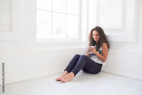 Portrait of glamour beautiful young woman with long curly hair in white room. Sit on floor with cup of drink. Positive stylish girl with smile on her face. © estradaanton