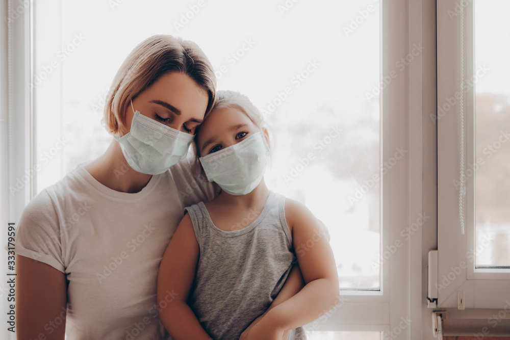 Family mom and daughter in medical mask. young woman and child little girl sitting by the window in protective masks against the virus     - obrazy, fototapety, plakaty 